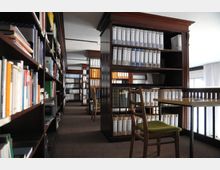 Innenraum einer Bibliothek mit hölzernen Bücherregalen, die dicht mit Büchern und Aktenordnern gefüllt sind. Im Vordergrund stehen ein Holztisch und ein Stuhl mit grünem Polster, während durch ein Fenster im Hintergrund Tageslicht einfällt.