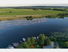 Luftaufnahme eines ruhigen Sees mit mehreren angelegten Booten an einem kleinen Steg. Im Vordergrund befinden sich eine Wiese mit Bäumen, einem Pavillon und roten Zelten; im Hintergrund erstrecken sich grüne Felder und Wälder.