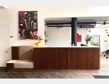 Modernes Empfangs- oder Thekenbereich mit einer holzverkleideten Front, einem beigefarbenen Sitzbereich an der linken Seite und einer offenen Wand mit Durchblick nach hinten. Ein abstraktes, farbenfrohes Kunstwerk hängt an der weißen Wand, mit einigen Pflanzen und einem Lampenschirm auf der Theke.