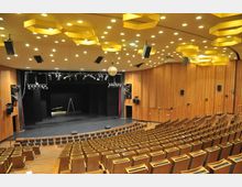Innenraum eines modernen Theatersaals mit ansteigenden Holzstühlen, einer breiten Bühne und Holzwänden. Die Decke ist mit gelben, hexagonalen Lampen und zahlreichen Einbauspots beleuchtet, während Bühnenscheinwerfer und eine Diskokugel von oben hängen.