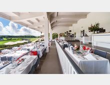 Überdachte Terrasse einer Rennbahn mit festlich gedeckten Tischen, weißen Tischdecken und gedeckten Stühlen; im Hintergrund ist die grüne Rennstrecke mit Sonnenschirmen zu sehen. Die Architektur ist hell, mit weiß gestrichenen Holzträgern und Rundbögen im Innenbereich.