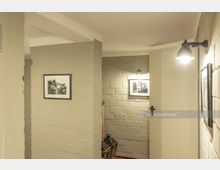 Flur in einem Gebäude mit schlicht verputzten Wänden und Mauerwerk aus hellem Ziegelstein, beleuchtet durch schwenkbare Wandlampen. An den Wänden hängen gerahmte Schwarz-Weiß-Fotografien, und eine Tür öffnet sich in einen angrenzenden Raum.