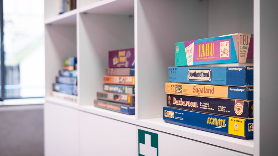 Regal mit mehreren Fächern, in denen Brettspiele wie "Tabu", "Scotland Yard" und "Activity" gestapelt sind; darunter befindet sich ein weißer Schrank mit einem grünen Erste-Hilfe-Kreuz. Im Hintergrund ist ein Fenster zu sehen.