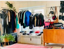 Raum mit hängender Kleidung auf Kleiderstangen, darunter Anzüge und glitzernde Abendgarderobe, sowie Taschen an der Wand. Im Vordergrund ein Holzmöbel, ein Topfpflanze und eine rote Teppichfläche, dahinter ein Fenster und ein Heizkörper mit Hüten darauf.