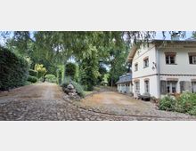 Landhausartige, weiß verputzte Gebäude mit Ziegelrahmungen um die Fenster und kleinem Vorplatz neben einer Kopfsteinpflaster-Zufahrt. Die Einfahrt ist von dichtem Grün, Hecken und großen Bäumen umgeben; am Rand liegen Findlinge und Bepflanzung.