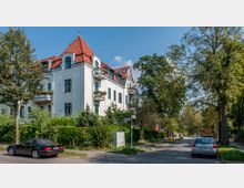 Eckansicht eines historischen Wohngebäudes mit hellem Putz, roten Dächern und kleinen Balkonen, umgeben von dichter Bepflanzung. Rechts verläuft eine baumbestandene Straße mit geparkten Autos.