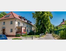 Mehrfamilienhaus in einer ruhigen Wohngegend mit hellem Putz und rotem Ziegeldach, daneben ein schmaler asphaltierter Weg, gesäumt von Bäumen und einem Zaun. Vor dem Gebäude befinden sich Blumenbeete, ein kleiner Balkon und ein geparkter blauer Wagen.