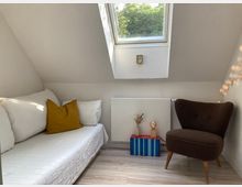 Gemütlicher Dachzimmerbereich mit einem weißen Tagesbett, dekoriert mit gelben und weißen Kissen. Rechts steht ein brauner Sessel, daneben eine kleine Dekoration aus Trockenblumen auf einem Bücherstapel unter einem schrägen Dachfenster, durch das Baumkronen zu sehen sind.