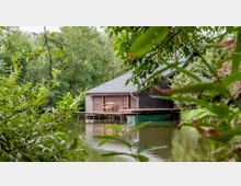Holzhaus mit grauem Satteldach, das auf Stelzen über einem ruhigen Gewässer gebaut ist, umgeben von dichtem Grün und Bäumen. Eine Holzterrasse mit einem Stuhl ist direkt am Wasser zu sehen.