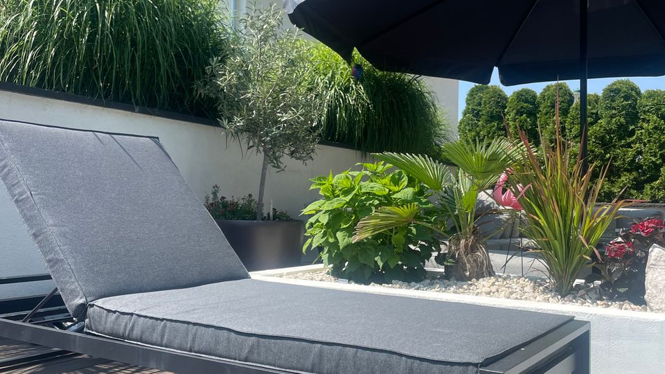 Sonnenliege auf einer Holzterrasse, umgeben von grünen Pflanzen in Blumenbeeten mit weißen Steinen. Im Hintergrund ein schwarzer Sonnenschirm und ein modernes Wohnhaus mit weißer Fassade.