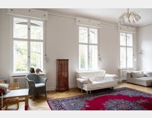 Helles Wohnzimmer mit hohen weißen Wänden und großen, doppelflügeligen Fenstern, die von Bäumen umgebenes Tageslicht hereinlassen. Der Raum ist mit einem roten Orientteppich, einer weißen Couch, einem Rattan-Sessel und einem verzierten Holzschrank eingerichtet, ergänzt durch Parkettboden und eine filigrane Deckenleuchte.