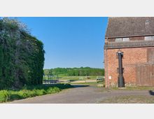 Backsteinbau mit Schornstein auf der rechten Seite und einer überwucherten Wand auf der linken Seite, dahinter eine grüne Wiese mit Holzzäunen, Hindernissen und einem Pferd; im Hintergrund ein dichter Wald unter klarem, blauem Himmel.