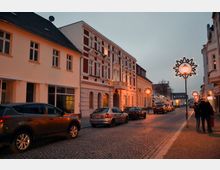 Straßenszene einer gepflasterten Straße mit historischen Wohnhäusern, darunter ein Gebäude mit roter und weißer Fassade. Entlang der Straße stehen geparkte Autos, und festliche Straßenlaternen mit sternförmigen Dekorationen beleuchten die Szene in der Dämmerung.