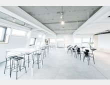 Modernes Konferenz- oder Schulungsraum mit hellen Wänden und großen, schräg angeordneten Fenstern, ausgestattet mit schwarzen Stühlen, kleinen Schreibpulten und weißen Stehtischen mit Hockern. Der Raum hat einen Betonboden und eine offene Decke mit sichtbaren Trägern und Beleuchtungsschienen.