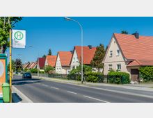 Straßenszene in einer Wohngegend mit einer Reihe von Einfamilienhäusern mit roten Satteldächern, gepflegten Gärten und Vorgärten, daneben eine Bushaltestelle mit einem gelben Haltestellenschild. Im Vordergrund verläuft eine asphaltierte Straße, auf der ein Auto zu sehen ist.