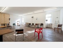 Ein einfacher Raum mit Holzfußboden und weißen Wänden, eingerichtet mit einer Mischung aus Stühlen unterschiedlicher Art, einem roten Plastikstuhl und ein paar kleinen Tischen. Im Hintergrund stehen eine Leiter, ein Heizkörper unter einem Fenster und zwei aufgestellte Lampen.