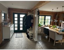 Eingangsbereich eines Hauses mit einer doppelflügeligen grünen Tür mit Glasfeldern und einem angrenzenden Essbereich. Der Raum besteht aus Holz- und Steinelementen, mit einem großen Holztisch, gepolsterten Stühlen und Hängelampen, sowie dekorativen Elementen wie einem Wanduhrengehäuse und Weinregalen.