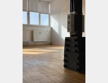 Leerer Raum mit hellem Holzboden, einer großen Fensterfront mit geschlossenen Jalousien und Heizkörpern an der Wand. Im Vordergrund ist ein massiver, schwarzer Metallträger mit Steckdosen und Kabeln sichtbar.