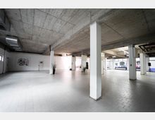 Großer, leerer Innenraum mit weiß gefliesten Böden, weißen Säulen und einer Decke aus Sichtbeton. Im Hintergrund befinden sich weiße Wände mit einem abstrakten Wandbild, einige Pflanzen in schwarzen Töpfen und große Fenster, die Tageslicht hereinlassen.