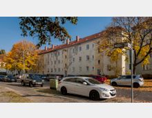 Wohngebäude aus mehreren Stockwerken mit heller Putzfassade und roten Ziegeldächern, umgeben von Herbstbäumen mit leuchtendem Laub. Auf der Straße im Vordergrund sind parkende Autos und ein Straßenschild mit der Aufschrift "Scheinerweg" sichtbar.