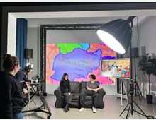 Ein Aufnahmestudio mit zwei Personen, die auf einem Sofa sitzen, während eine Kamera und eine große Studiolampe auf sie gerichtet sind. Im Hintergrund ist eine große Projektion einer Afghanistan-Karte sowie ein Bildschirm mit einem Bild einer Gruppe von Menschen zu sehen.