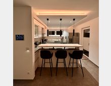 Moderne Küche mit zentraler Kochinsel, darüber drei hängende schwarze Pendelleuchten. Hinter der Insel befinden sich Einbauküchenschränke, ein Edelstahlkühlschrank und ein Fenster; der Raum ist mit warmem indirekten Licht beleuchtet.