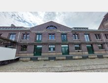 Backsteinfassade einer historischen Lagerhalle mit der Aufschrift „Lagerhalle I.“ über dem Eingangsbereich. Die grüne Doppeltür und Fensterrahmen aus Metall sowie das gepflasterte Gelände davor sind deutlich sichtbar; im Vordergrund steht links ein weißer Lieferwagen.