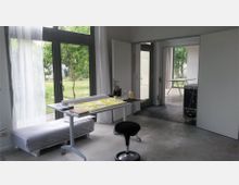 Heller Arbeitsraum mit großen Glasfronten, die den Blick in einen grünen Garten ermöglichen. Im Vordergrund steht ein Schreibtisch mit Malutensilien, daneben ein Hocker und eine Liege, im Hintergrund ein offener Durchgang zu einem weiteren Raum.