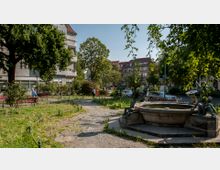 Kleiner Stadtpark mit einem Brunnen, umgeben von Grünflächen, Bäumen und Bänken. Im Hintergrund sind Wohnhäuser sowie parkende Autos entlang der Straße zu sehen.