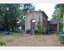 Backsteinbau mit dekorativer Fassade und zentralem Rundbogenportal, flankiert von modernen, flach konstruierten Anbauten mit großen Fenstern. Das Gebäude steht in einem grasbewachsenen Bereich, umgeben von hohen Bäumen; im Vordergrund befinden sich blaue Gartenstühle und ein Tisch.