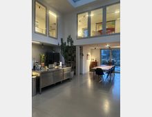 Moderner Wohnraum mit hohem, offenem Bereich und einer Galerieebene, die durch Fenster sichtbar ist. Im unteren Bereich stehen eine Edelstahlküche, ein großer Esstisch mit schwarzen Stühlen und dekorative Pflanzen, darunter ein großer Kaktus; im Hintergrund ist ein großes Fenster mit Blick nach draußen.