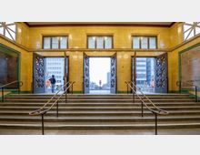 Eingangsbereich einer U-Bahn-Station mit gelb gefliesten Wänden, Treppen mit Metallgeländern, geöffneten Doppeltüren und Blick nach draußen auf städtische Gebäude und Fußgänger.