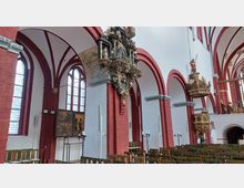 Innenraum einer gotischen Kirche mit roten Backsteinpfeilern und Spitzbögen, weiß gestrichenen Wänden und großen, hohen Fenstern mit Glasmalereien. Auffällig sind die detaillierten, historischen Schnitzwerke an den Pfeilern und die reich verzierte Kanzel auf der rechten Seite, umgeben von Reihen hölzerner Kirchenbänke.