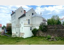 Weiße historische Gebäude mit Treppengiebeln und leicht abgeblätterter Fassade, daneben eine alte Steinmauer. Im Vordergrund liegt eine Grünfläche mit Wildblumen, und im Hintergrund sind Bäume sowie weitere Gebäude sichtbar.