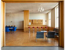 Ein heller, holzvertäfelter Raum mit Parkettboden, einer Theke mit roten Barhockern und einer Sitzgruppe aus blauen Sesseln und Tisch. An der linken Seite steht ein Esstisch mit blauen Stühlen, während große Fenster auf der rechten Seite Tageslicht hereinlassen.