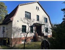 Zweistöckiges Einfamilienhaus mit weißer Putzfassade, grün gerahmten Fenstern und einem roten Satteldach. Der Eingangsbereich ist über eine kleine Treppe mit gemauerten Wänden erreichbar, umgeben von einem Garten mit Bäumen und Sträuchern.