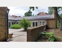 Backsteingebäude mit Flachdach in einem von Bäumen umgebenen Außenbereich. Vor dem Gebäude befindet sich eine gepflasterte Terrasse mit Pflanzenbeeten und einer niedrigen Backsteinmauer.