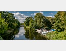 Flusslandschaft mit dicht bewaldeten Ufern und mehreren kleinen Booten, die an einem Steg am rechten Ufer festgemacht sind. Im Hintergrund ist eine Brücke zu sehen, während der Himmel blau ist und von weißen Wolken durchzogen wird.