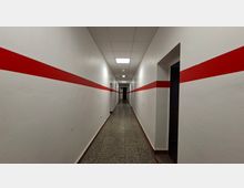 Langer, schmaler Innenflur mit weißen Wänden und einem durchgehenden roten Querstreifen auf beiden Seiten. Grauer Steinboden, abgehängte Decke mit rechteckigen Deckenleuchten und mehrere Türöffnungen entlang des Gangs.