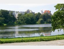 Ein ruhiger Park mit einem See im Vordergrund, umgeben von grünen Bäumen. Im Hintergrund sind Gebäude mit unterschiedlicher Architektur, darunter ein Haus mit rotem Dach und ein Turm mit Fahne, zu sehen.