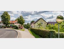 Straßenszene in einer Vorstadt mit einer asphaltierten Straße, Gehweg, und Einfamilienhäusern mit roten und dunklen Dächern. Im Vordergrund sind Büsche, Bäume und ein Verkehrsspiegel am Straßenrand sichtbar, während der Himmel mit Wolken bedeckt ist.