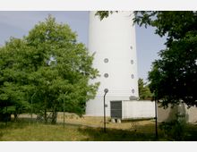 Ein hoher, weißer Turm mit runden Fenstern steht hinter einem Maschendrahtzaun, umgeben von grünen Bäumen. Am Fuß des Turms befinden sich technische Einrichtungen und eine eingezäunte Wiese.