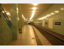 Unterirdischer Bahnhof mit gekachelten Wänden in hellgrüner Farbe, beleuchtet durch eine Reihe von Deckenlampen; der Bahnsteig verläuft entlang gebogener Gleise. Auf den Wänden ist der Stationsname "Alexanderplatz" mehrfach angebracht.