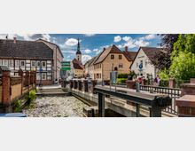 Historische Altstadt mit Fachwerk- und Ziegelgebäuden sowie einer kleinen Brücke über einen flachen Wasserlauf; im Hintergrund ein Kirchturm und ein blauer Himmel mit vereinzelten Wolken.