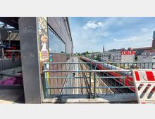 Bahnhofsbereich mit Gleisen und vorbeifahrendem Zug, umgeben von modernen Gebäuden, darunter eine mit Werbeplakaten und Graffiti versehene Wand. Im Hintergrund eine Baustelle, weitere Gebäude, und ein Kirchturm unter klarem Himmel sichtbar.