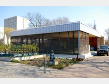 Modernes, einstöckiges Gebäude mit einer Fassade aus horizontalen Lamellen, das von einem kleinen Garten mit Blumenbeeten und einer Skulptur in der Mitte umgeben ist. Im Hintergrund sind mehrstöckige Wohnhäuser und kahle Bäume zu sehen.