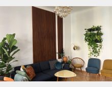 Gemütliches Wohnzimmer mit modernem Design: Dunkelblaue Couch mit bunten Kissen, daneben ein heller Couchtisch. An der Wand hängen vertikale Holzpaneele und eine Pflanzenhalterung mit rankenden Pflanzen. Der Raum ist mit Stühlen im Retro-Stil, einem goldenen Bogenlampen und Holzfußboden eingerichtet.