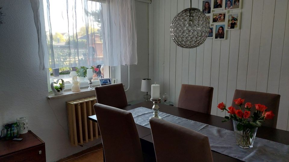 Heller Essbereich mit dunklem Esstisch und vier gepolsterten Stühlen, dekoriert mit Tischläufer, Kerzenhalter und einer Glasvase mit roten Blumen. An der Wand sind weiße Holzpaneele und eine Fotocollage zu sehen; daneben ein Fenster mit transparenten Vorhängen, Fensterbank-Deko und Heizkörper darunter sowie eine runde Hängelampe aus Metallgeflecht.