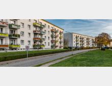 Mehrstöckige Wohngebäude mit farbigen Balkonfassaden stehen entlang einer gepflasterten Straße. Im Vordergrund befinden sich eine Grünfläche, gepflegte Hecken und ein Verkehrsschild mit einer Tempo-30-Zone.