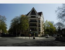 Mehrstöckiges Wohngebäude an einer Straßenecke, mit spitzem Giebeldach und Balkonen. Umgeben von Bäumen, geparkten Autos und Gehwegen, lädt ein kleiner Bereich mit Sonnenschirmen und Sitzgelegenheiten vor dem Gebäude zum Verweilen ein.